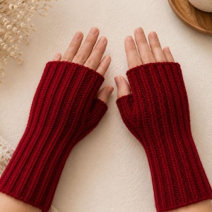 May include: A pair of deep red fingerless gloves featuring a vertical ribbed knit pattern. The gloves extend from the knuckles down to the mid-forearm, leaving the fingers and thumbs exposed. The knit texture is consistent throughout the garment.