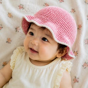 Puede incluir: Un sombrero de pescador tejido a ganchillo en color rosa con un borde de color crema. El sombrero presenta un patrón de punto acanalado y texturizado en toda la copa y un ala ligeramente acampanada. El diseño es sencillo y destaca el tejido.