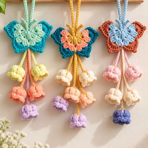 May include: Three hanging crochet butterfly ornaments with dangling bell-shaped flowers. The left butterfly is mint and teal with yellow, peach, and purple flowers. The center is yellow and teal with white, peach, and purple flowers. The right is blue and brown with pink, white, and blue flowers.