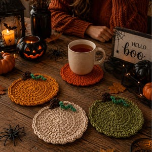 May include: Three round, crocheted coasters shaped like pumpkins in orange, beige, and olive green. Each coaster features a small brown stem and a green vine detail. The coasters are arranged on a wooden surface alongside a mug and decor.