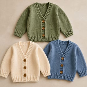 Puede incluir: Tres cárdigans de punto de manga larga en verde oliva, crema y azul polvoriento. Cada cárdigan presenta un cuello en V, puños y dobladillo acanalados, y tres botones de madera marrón en la parte central delantera. La textura es uniforme.