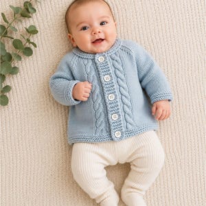 May include: A light blue long-sleeve knit cardigan featuring a cable knit pattern, a round neckline, and five white buttons down the front, paired with cream-coloured ribbed knit footed leggings. The garment is worn by a smiling infant.