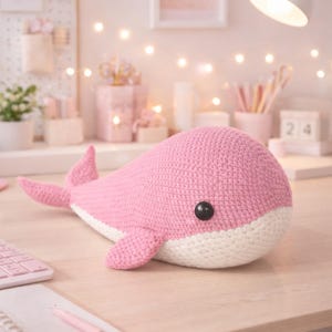 Pode incluir: Uma baleia rosa de crochê feita à mão com barriga branca e olhos de botão pretos repousa sobre uma superfície de madeira clara. O fundo inclui uma luminária de mesa e luzes de fada. Um brinquedo macio para o quarto de uma criança.
