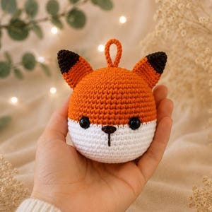 Könnte beinhalten: Ein rundes, gehäkeltes Fuchsornament mit einem leuchtend orangefarbenen Oberkopf und einem weißen Gesichtsbereich. Es besitzt zwei spitze Ohren mit dunkelbraunen Spitzen, zwei kleine schwarze Perlenaugen, eine kleine schwarze Nase und eine Schlaufe.
