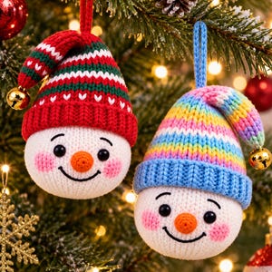 Puede incluir: Dos adornos de muñeco de nieve tejidos y redondos con caras bordadas, narices naranjas y mejillas rosadas. Uno lleva un gorro de rayas rojas, verdes y blancas, el otro un gorro de rayas arcoíris. Ambos gorros tienen un cascabel dorado en la punta.