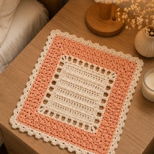 May include: A rectangular crochet mat featuring a central cream-colored panel with horizontal textured rows, surrounded by a wide border of peach-colored yarn and finished with a delicate cream-colored scalloped edge along the entire perimeter.