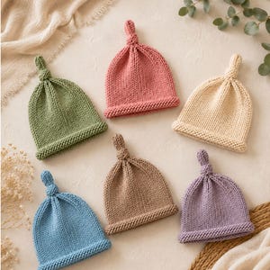 Puede incluir: Seis gorros tejidos con un nudo en la parte superior y borde enrollado, dispuestos en un patrón circular. Los gorros son de color verde salvia, rosa viejo, crema, azul claro, beige y lavanda, con una textura de punto jersey uniforme.