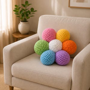 May include: A flower-shaped pillow composed of eight crocheted spheres in various colors: pink, yellow, orange, green, blue, purple, and white. The pillow rests on a beige armchair, creating a vibrant accent in a neutral-toned living space.