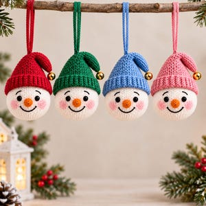 Könnte beinhalten: Vier runde, gestrickte Schneemann-Anhänger, die an einem Ast hängen. Jedes Gesicht ist weiß mit schwarzen Augen, einer kleinen orangefarbenen Nase und rosa Wangen. Sie tragen gestrickte Mützen in Rot, Grün, Blau und Pink mit einer Goldglocke.