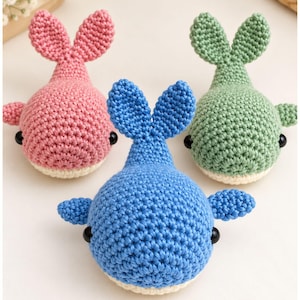 May include: Three small, handmade crochet whale toys in pink, blue, and green. Each whale features a rounded body, a vertical tail fin, two side fins, and two small black bead eyes. The underside of each whale is a cream-coloured yarn.