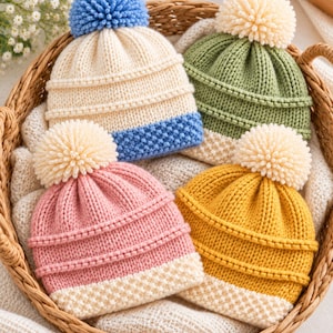 May include: Four knitted beanies with pom-poms, each featuring a textured ribbed crown and a seed-stitch brim. The hats are colored in cream with a blue brim, sage green with a cream pom-pom, dusty pink with a cream brim, and mustard yellow with a cream brim.