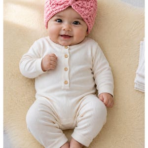 May include: A cream-colored, long-sleeved, ribbed cotton romper featuring a button-down front placket with four small wooden buttons, paired with a textured, dusty pink crocheted turban-style hat with a central knot detail.