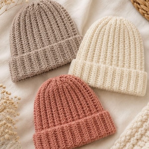 Pode incluir: Três gorros de malha em cores sólidas: um taupe, um creme e um rosa antigo. Cada gorro apresenta uma textura canelada vertical e uma dobra larga na base. Os gorros estão dispostos em grupo, destacando a construção consistente do fio e a paleta de cores suaves e distintas de cada peça individual.