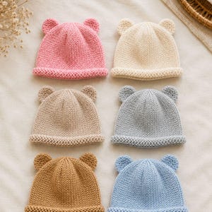 May include: A collection of six knitted beanies, each featuring two small rounded ears on top. The hats are presented in a variety of solid colors including pink, cream, beige, light gray, tan, and light blue, all with a rolled brim edge.