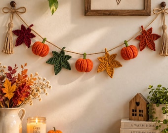 May include: A decorative garland featuring crocheted orange pumpkins and multi-colored autumn leaves strung on a jute twine rope with beige tassels at each end. The garland is displayed against a wall, hanging between two wooden knobs.