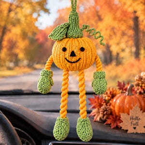 Könnte beinhalten: Ein gestricktes Kürbis-Autoornament mit einem leuchtend orangefarbenen runden Körper, einem schwarzen gestickten Gesicht, einem grünen Blatt und einer Ranke. Es hat lange gelbe Seilarme und -beine mit grünen gestrickten Fäustlingen und Stiefeln.