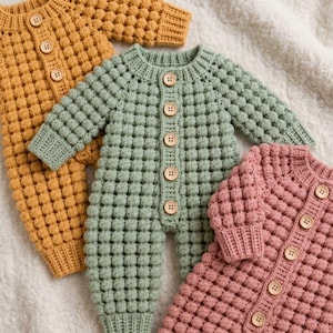 May include: Three long-sleeve knitted rompers featuring a textured bobble stitch pattern. Each romper has a round neckline, ribbed cuffs, and a vertical front placket with five light brown wooden buttons. The rompers are mustard yellow, sage green, and dusty pink.