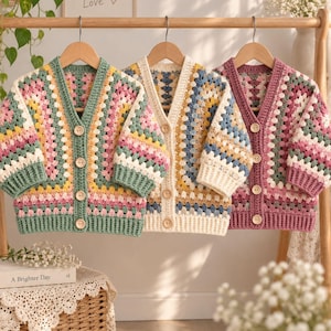May include: Three crochet cardigans hanging on wooden hangers. The left cardigan features green, pink, and yellow yarn. The middle cardigan is cream, blue, and yellow. The right cardigan is pink, green, and white. Each has four light wooden buttons.