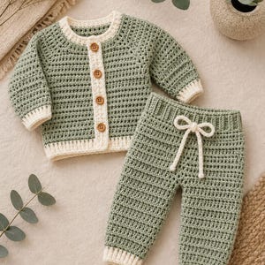 Può includere: Un completo in due pezzi all'uncinetto color verde salvia e crema, composto da un cardigan a maniche lunghe e pantaloni abbinati. Il cardigan presenta un'abbottonatura color crema con quattro bottoni marroni. I pantaloni hanno una coulisse.
