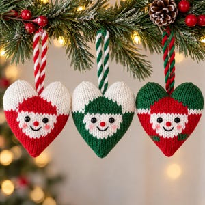 May include: Three heart-shaped knitted ornaments featuring smiling faces with black bead eyes, red noses, and pink cheeks. Each ornament has a twisted rope loop for hanging. The designs include red and white, green and white, and red and green.
