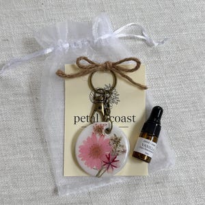 May include: A round white resin keychain featuring embedded pink and dried flowers, attached to a bronze-toned metal clasp. It is displayed on a cream card labeled petal coast with a brown twine bow, alongside a 3ml amber glass bottle of vanilla oil.