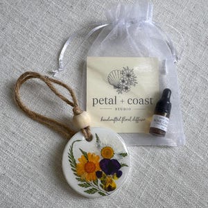 May include: A round white ceramic diffuser disc decorated with pressed yellow and purple flowers and green ferns, suspended by a jute twine cord with a wooden bead. Includes a small brown glass dropper bottle and a white organza bag with a card.