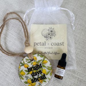 May include: A round ceramic diffuser with a lemon pattern and the text "bright days ahead" hanging from a jute cord with a wooden bead. Includes a 3ml amber glass bottle of lemon essential oil and a branded card in a white organza bag.