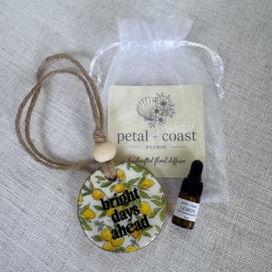 May include: A round ceramic diffuser with a lemon pattern and the text bright days ahead, featuring a jute hanging cord and a wooden bead. Included is a small amber glass bottle of lemon essential oil and a white organza bag with a petal + coast studio card.