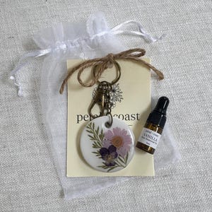 May include: A round white ceramic keychain featuring pressed purple and pink flowers with green leaves, attached to a bronze metal clasp. It is displayed on a branded card with a small 3ml amber glass bottle of vanilla essential oil and a white organza bag.