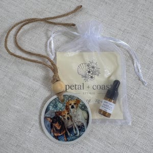 May include: A round ceramic hanging ornament featuring a printed image of three small dogs, suspended by a brown jute cord with a wooden bead. Included is a small amber glass dropper bottle labeled Vanilla and a white organza drawstring bag.