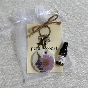 May include: A round white ceramic keychain featuring a pressed purple flower and green fern design, attached to a bronze-toned metal ring. Included is a 3mL amber glass bottle labeled Petal Coast Vanilla Pure Essential Oil on a card.