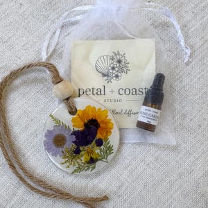 May include: A round white diffuser disc featuring embedded pressed yellow, purple, and green flowers, suspended by a jute cord with a wooden bead. Included is a small brown glass bottle of Rose Garden essential oil and a white organza bag.