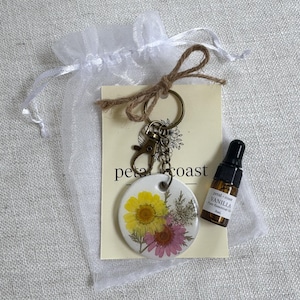 May include: A round white keychain featuring pressed yellow and pink flowers, attached to a bronze-toned metal ring and clasp. It is displayed on a branded card labeled petal + coast, accompanied by a 3ml amber glass bottle of vanilla essential oil.