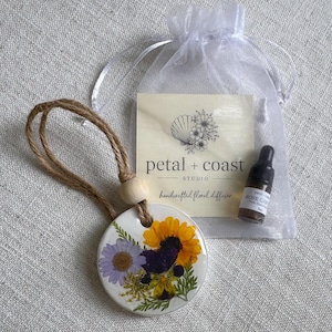 May include: A round white ceramic diffuser ornament decorated with pressed yellow, purple, and green flowers, hanging from a jute twine cord with a wooden bead. Includes a small amber glass dropper bottle labeled Rose Garden and a white organza bag.