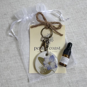 May include: A round white ceramic diffuser keychain featuring a pressed blue flower and green leaves, attached to a bronze metal clasp. It includes a small 5ml amber glass bottle of vanilla essential oil and a branded card in a white organza bag.