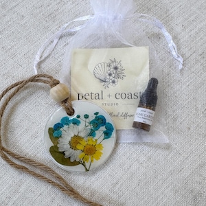 May include: A white circular ceramic diffuser pendant featuring pressed blue, white, and yellow flowers, suspended by a jute cord with a wooden bead. Included is a small amber glass bottle of Rose Garden essential oil and a white organza bag.