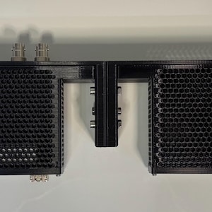 May include: A black, rectangular audio component with a series of perforated, honeycomb-patterned enclosures. The device has multiple connectors and is mounted on a white wall. The design is modern and industrial.