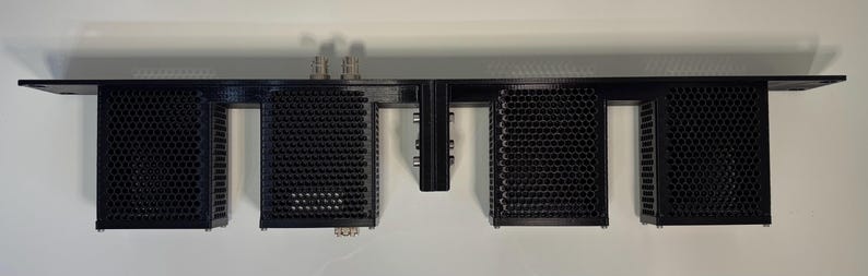 May include: A black, rectangular audio component with a series of perforated, honeycomb-patterned enclosures. The device has multiple connectors and is mounted on a white wall. The design is modern and industrial.