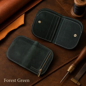 May include: A forest green leather wallet featuring rounded corners, white contrast stitching, and a gold-toned metal zipper on the exterior. The interior displays multiple card slots and a gold-toned snap button closure for secure storage.