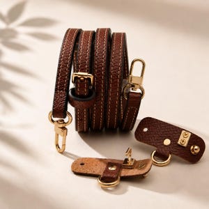 May include: A set of brown leather bag straps and hardware. The straps are rolled up, showing white stitching and gold-colored metal clasps. Additional leather pieces with gold hardware are also displayed.
