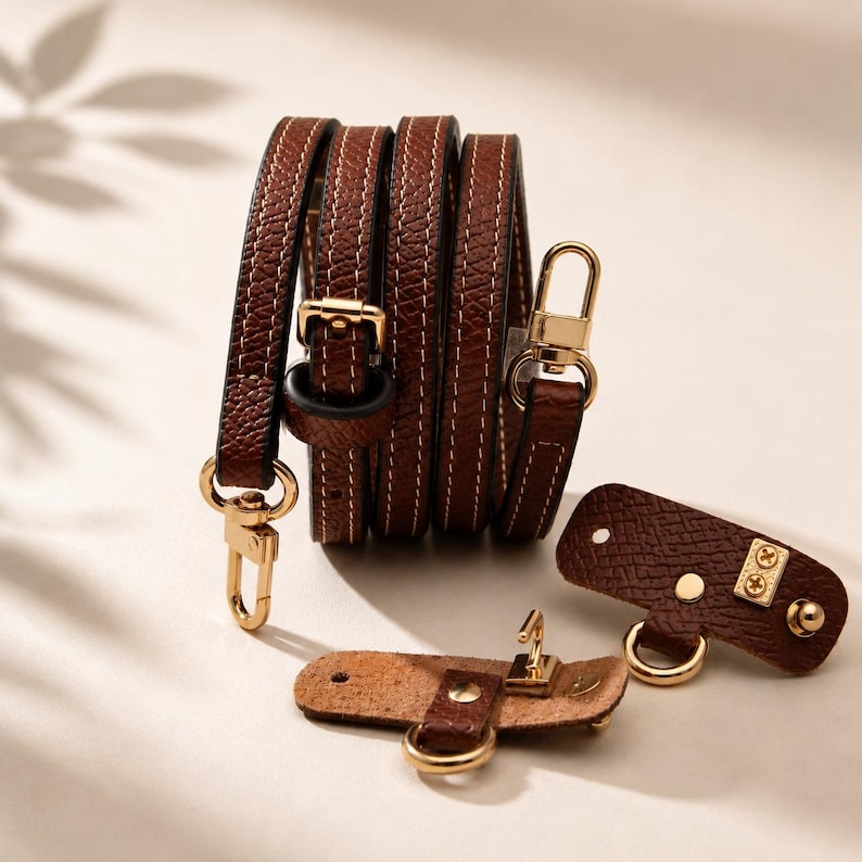 Puede incluir: Una correa de bolso de cuero texturizado marr&oacute;n oscuro con costuras blancas en contraste y herrajes de metal dorado, que incluye una hebilla y mosquetones giratorios, acompa&ntilde;ada de dos peque&ntilde;as pesta&ntilde;as de fijaci&oacute;n a juego con anillas.
