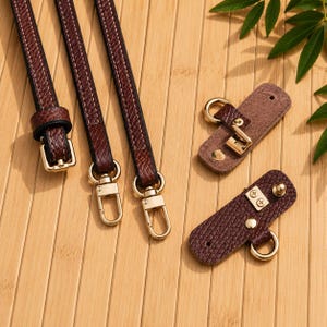 Puede incluir: Tres correas texturizadas de color marr&oacute;n oscuro con hebillas y mosquetones giratorios de metal dorado, junto con dos leng&uuml;etas rectangulares de piel sint&eacute;tica texturizada marr&oacute;n oscuro con anillos, tachuelas y tornillos dorados.