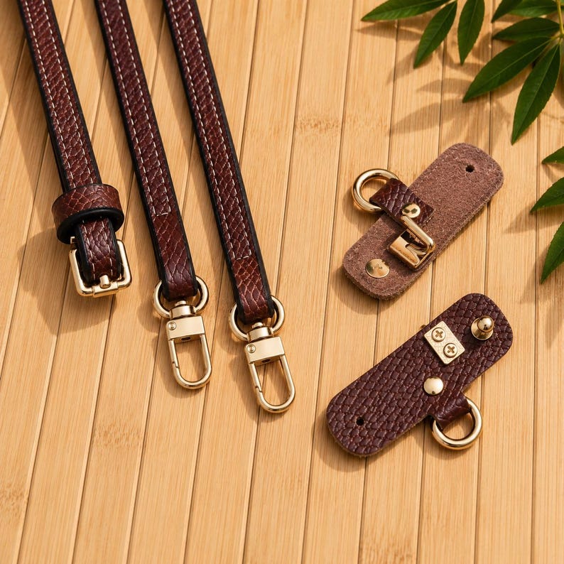 Puede incluir: Tres correas texturizadas de color marr&oacute;n oscuro con hebillas y mosquetones giratorios de metal dorado, junto con dos leng&uuml;etas rectangulares de piel sint&eacute;tica texturizada marr&oacute;n oscuro con anillos, tachuelas y tornillos dorados.