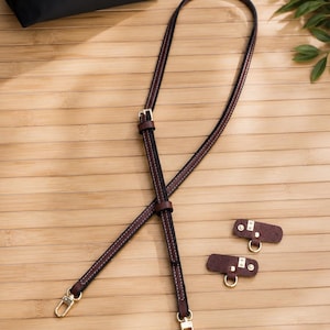Puede incluir: Una correa cruzada de cuero marr&oacute;n oscuro con cierres de mosquet&oacute;n de metal dorado y una hebilla ajustable, acompa&ntilde;ada de dos pesta&ntilde;as de fijaci&oacute;n de cuero marr&oacute;n a juego con anillos de metal dorado, junto a un bolso negro con solapa marr&oacute;n.