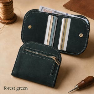 May include: A forest green leather wallet featuring a zippered exterior pocket and an open interior with multiple card slots. The wallet has rounded edges, visible white stitching, and gold-toned metal snap closures. A bill is tucked inside.
