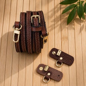 Puede incluir: Una correa de bolso de cuero texturizado marr&oacute;n oscuro enrollada con costuras blancas y mosquetones de metal dorado, acompa&ntilde;ada de dos leng&uuml;etas de fijaci&oacute;n de cuero marr&oacute;n a juego con anillas en D y botones a presi&oacute;n dorados.