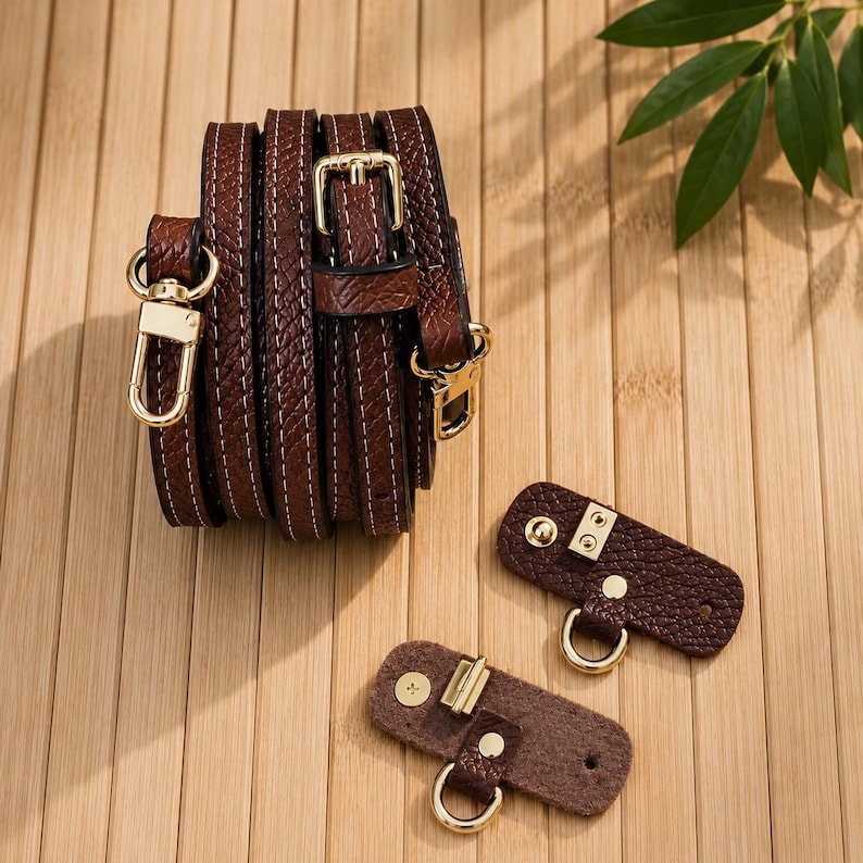 Puede incluir: Una correa de bolso de cuero texturizado marr&oacute;n oscuro enrollada con costuras blancas y mosquetones de metal dorado, acompa&ntilde;ada de dos leng&uuml;etas de fijaci&oacute;n de cuero marr&oacute;n a juego con anillas en D y botones a presi&oacute;n dorados.