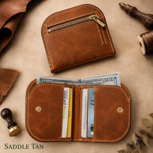 May include: A saddle tan leather wallet featuring a curved top edge, a zippered exterior pocket with a metallic zipper, and an open interior with multiple card slots and a bill compartment containing a one hundred dollar bill.