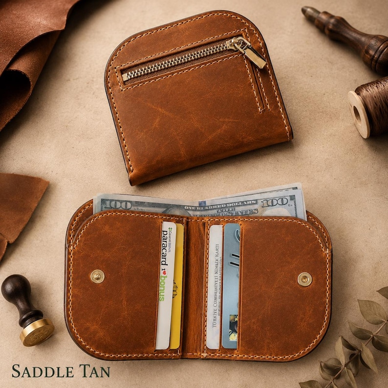 May include: A saddle tan leather wallet featuring a curved top edge, a zippered exterior pocket with a metallic zipper, and an open interior with multiple card slots and a bill compartment containing a one hundred dollar bill.