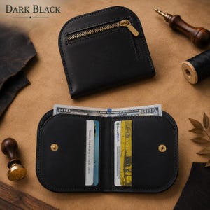 May include: A black leather wallet with a rounded top edge and a gold-tone metal zipper pocket on the exterior. The interior features card slots containing identification cards and a compartment holding a one hundred dollar bill.